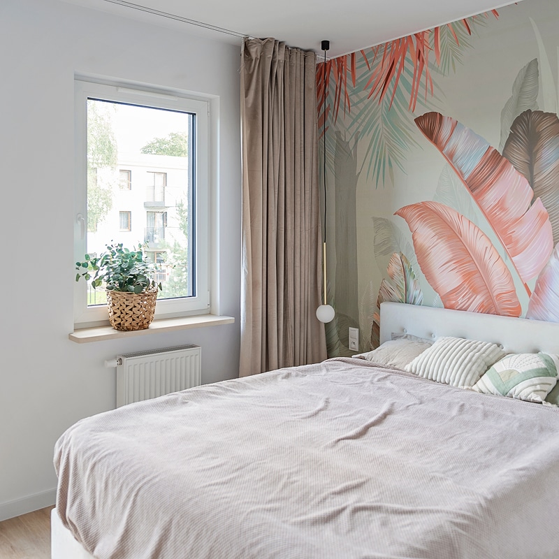 Chambre moderne lumineuse avec lit fait, papier peint tropical à grandes feuilles roses, fenêtre, plante tressée, rideaux.