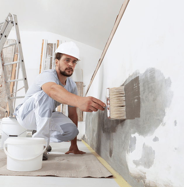 Rénovation Murale : Préparation avec Primaire par un Pro Un ouvrier en casque applique un primaire gris sur un mur avec une brosse lors d'une rénovation, avec échelle et seaux.