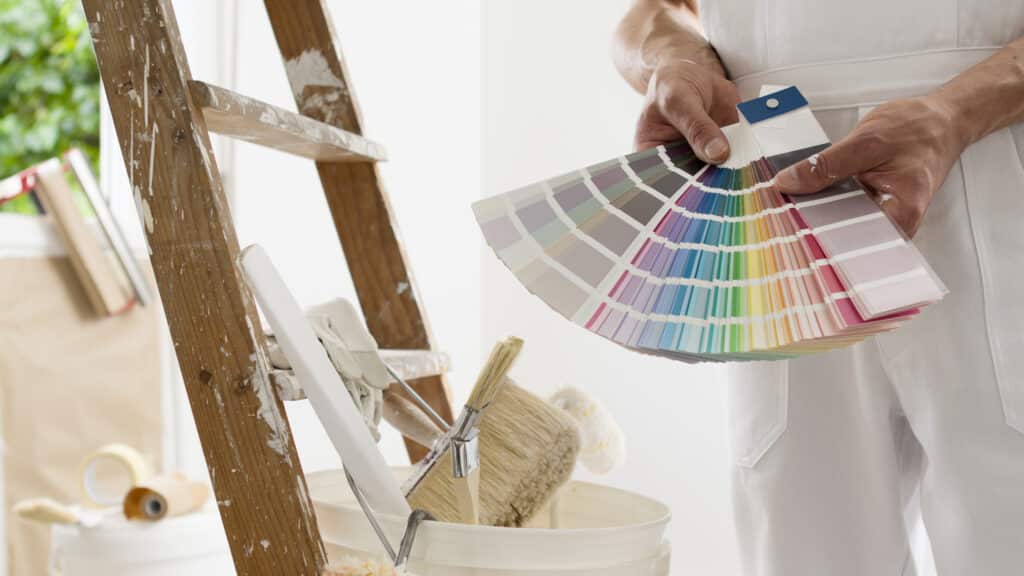 Peinture : Choisir ses couleurs avec un nuancier de peintre Peintre en salopette blanche tenant un nuancier de couleurs près d'une échelle tachée de peinture et d'outils de rénovation.
