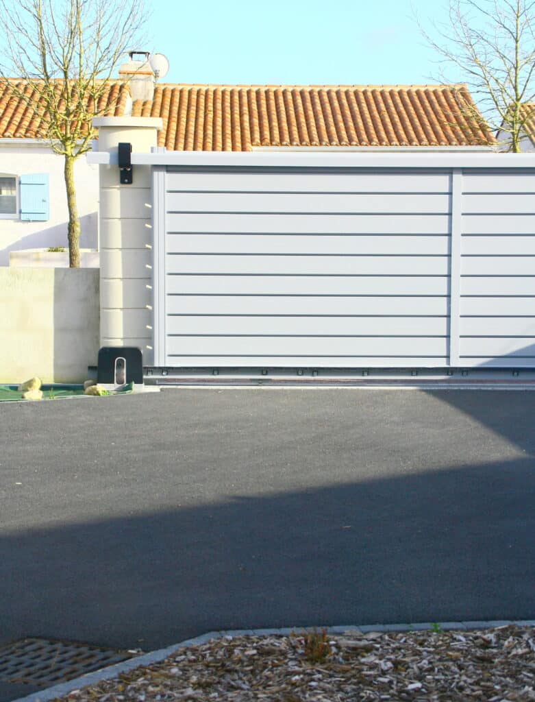 Portail coulissant blanc motorisé pour maison moderne Portail coulissant motorisé blanc devant une maison au toit de tuiles orange et ciel bleu. Allée goudronnée ensoleillée.