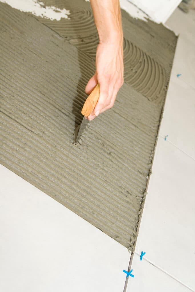 Main appliquant du mortier-colle gris en sillons avec une spatule pour la pose de carrelage blanc. Croisillons bleus visibles.