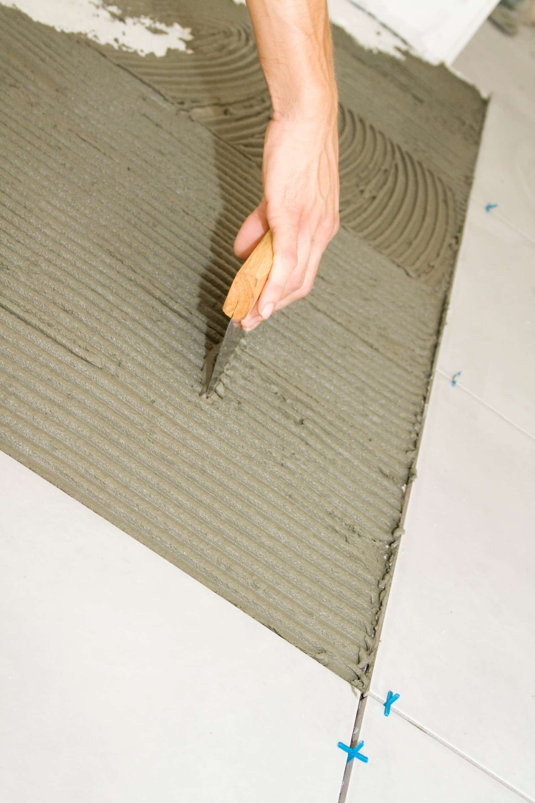 Main appliquant du mortier-colle gris en sillons avec une spatule pour la pose de carrelage blanc. Croisillons bleus visibles.